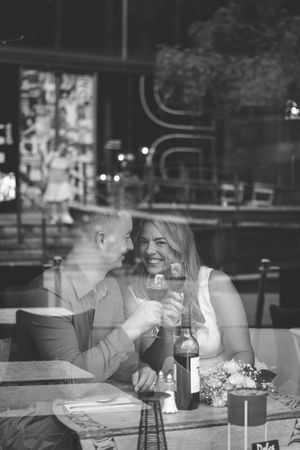 Black and white photo of a couple toasting wine glasses at a restaurant table, smiling and enjoying a romantic dinner date.