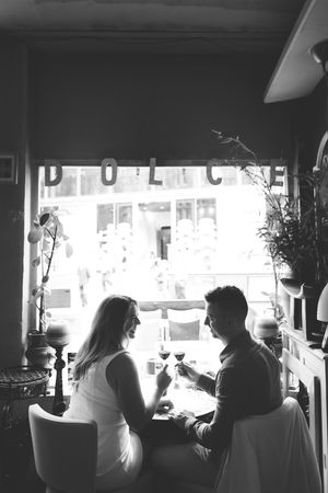 Black and white photo of a couple toasting wine glasses at a cafe table by a window with DOLCE sign.