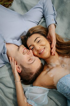A couple lying on a blanket outdoors sharing a tender moment while smiling and touching each other's faces.
