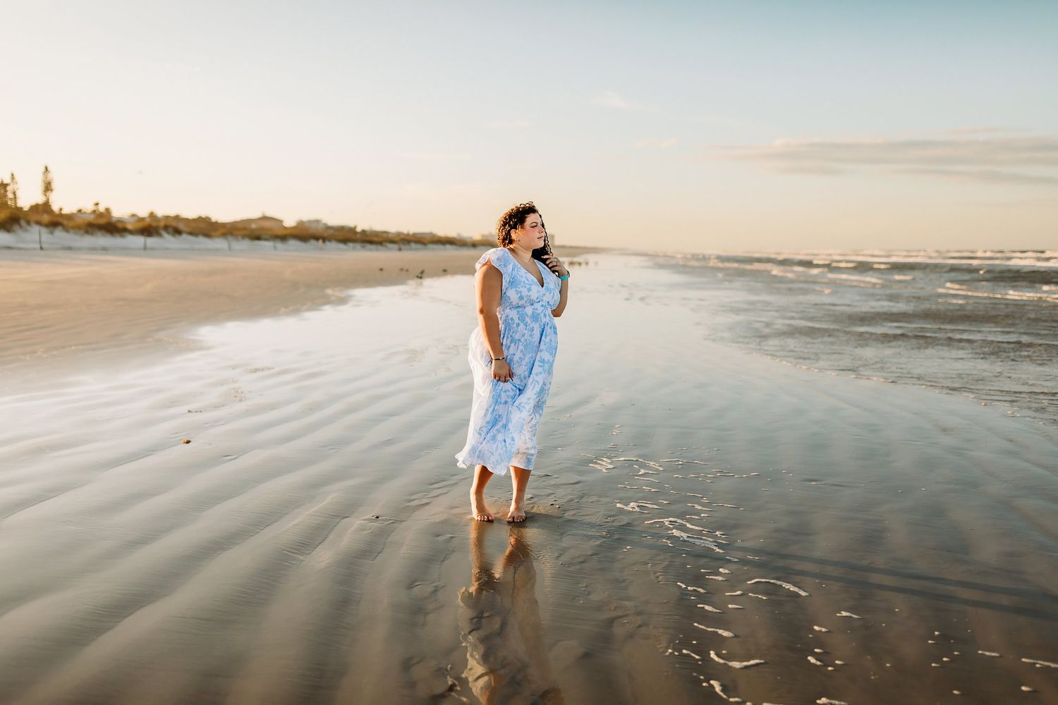 A person walks along a serene beach shoreline at sunset with calm waves rolling onto the sand.