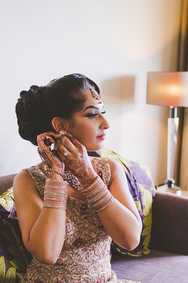 Traditional Indian bride wearing an embellished gown and henna adorned hands puts on jewelry in soft lighting.