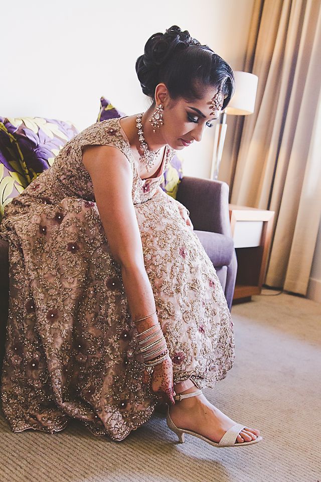 A bride in an embellished blush lehenga adjusts her silver sandal while getting ready in a hotel room.