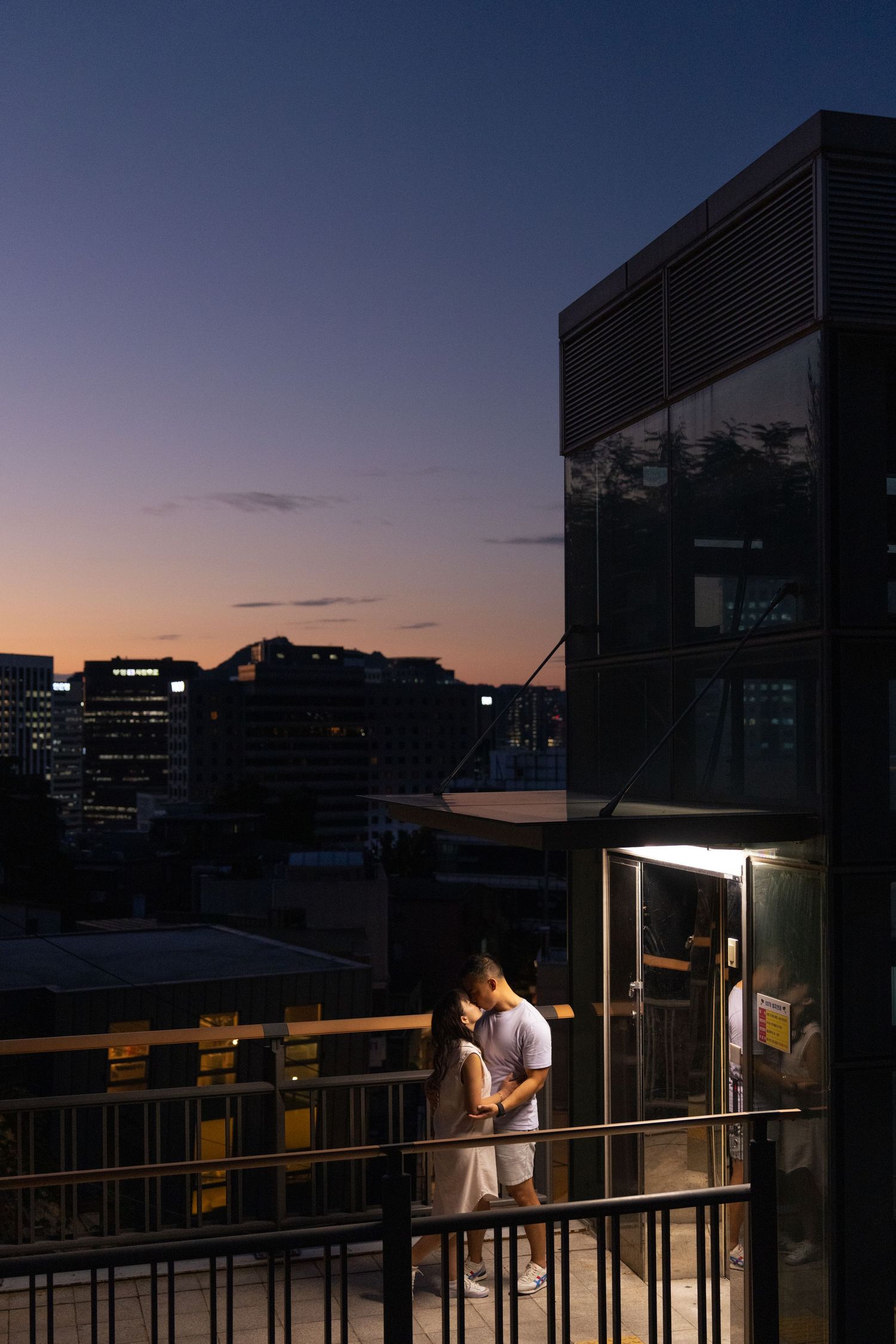 Person stands on a balcony at dusk with city buildings silhouetted against the darkening sky.