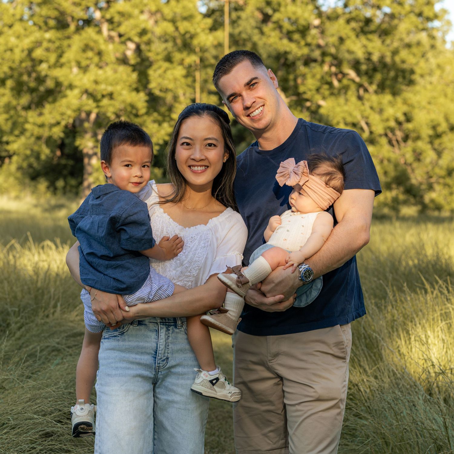 A happy young family of four poses outdoors in a grassy field, parents holding a toddler and newborn baby.