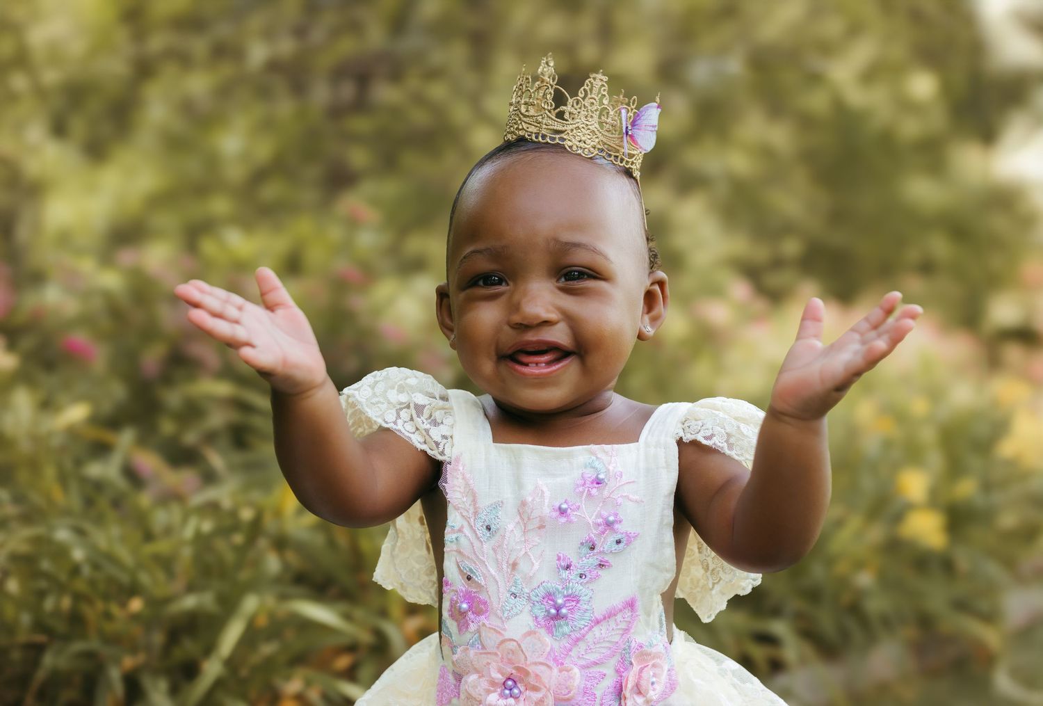 A baby wearing a floral dress and crown smiles joyfully while raising their arms in an outdoor setting.