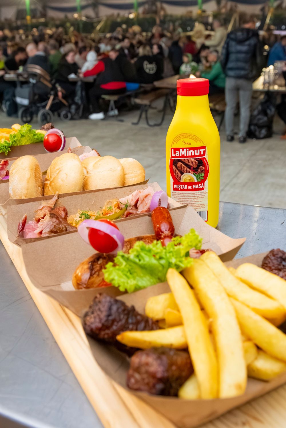 Fast food meal with french fries, sandwiches and yellow Smirnoff bottle served on wooden board at crowded outdoor venue.