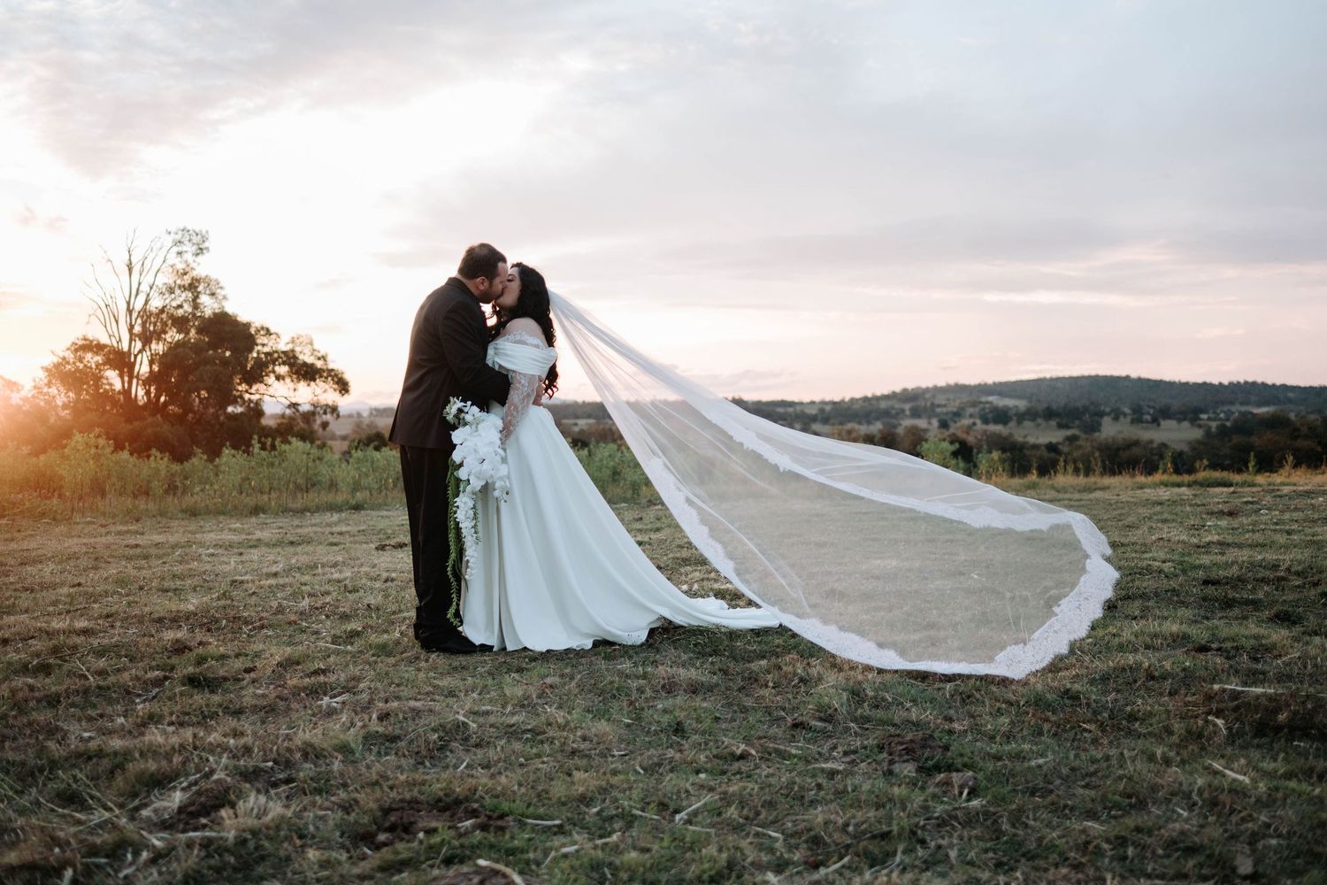 A romantic couple in wedding attire embraces in a scenic field at sunset as a long veil flows dramatically in the wind.