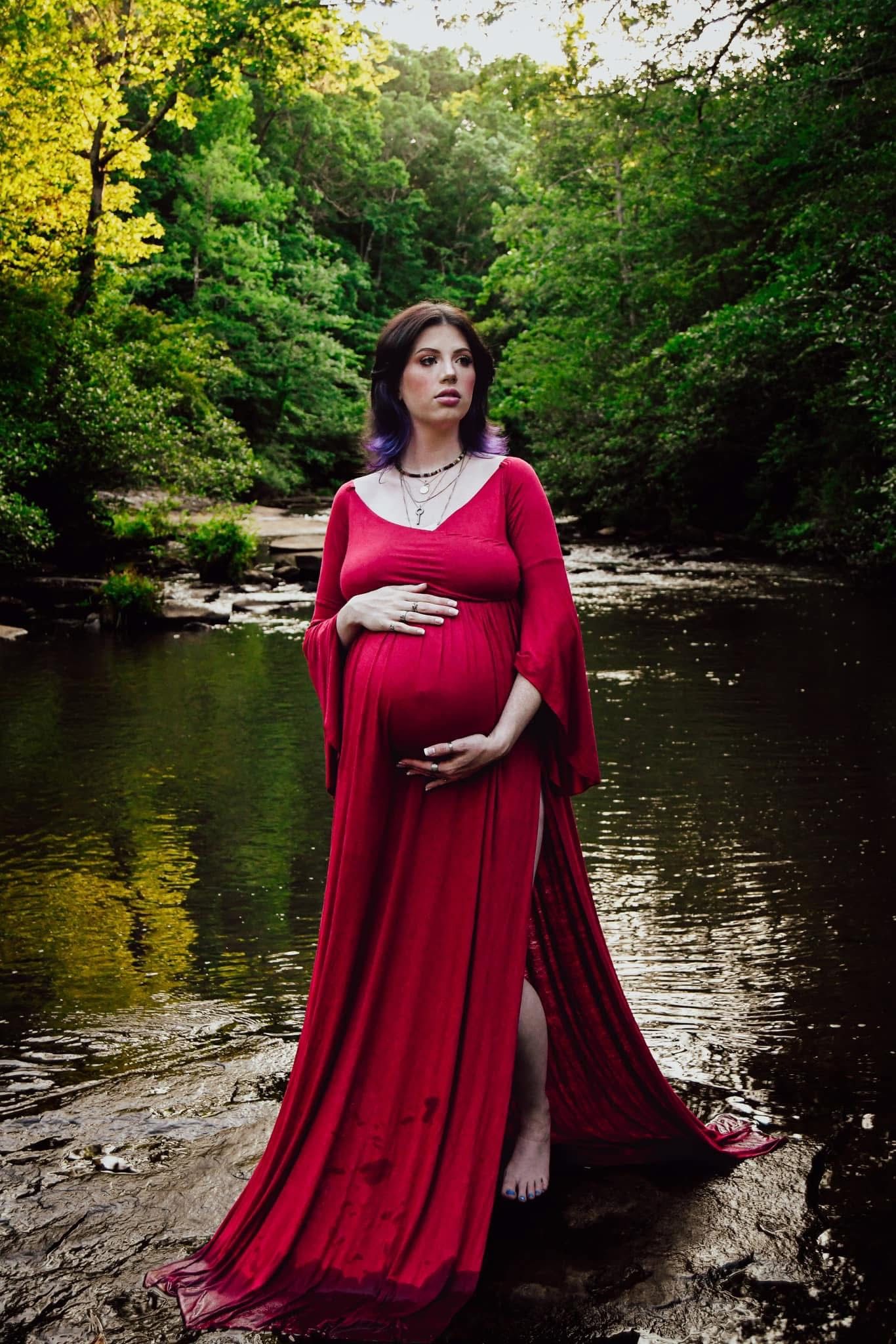 Mom to be in flowing red dress poses dramatically by wooded stream.
