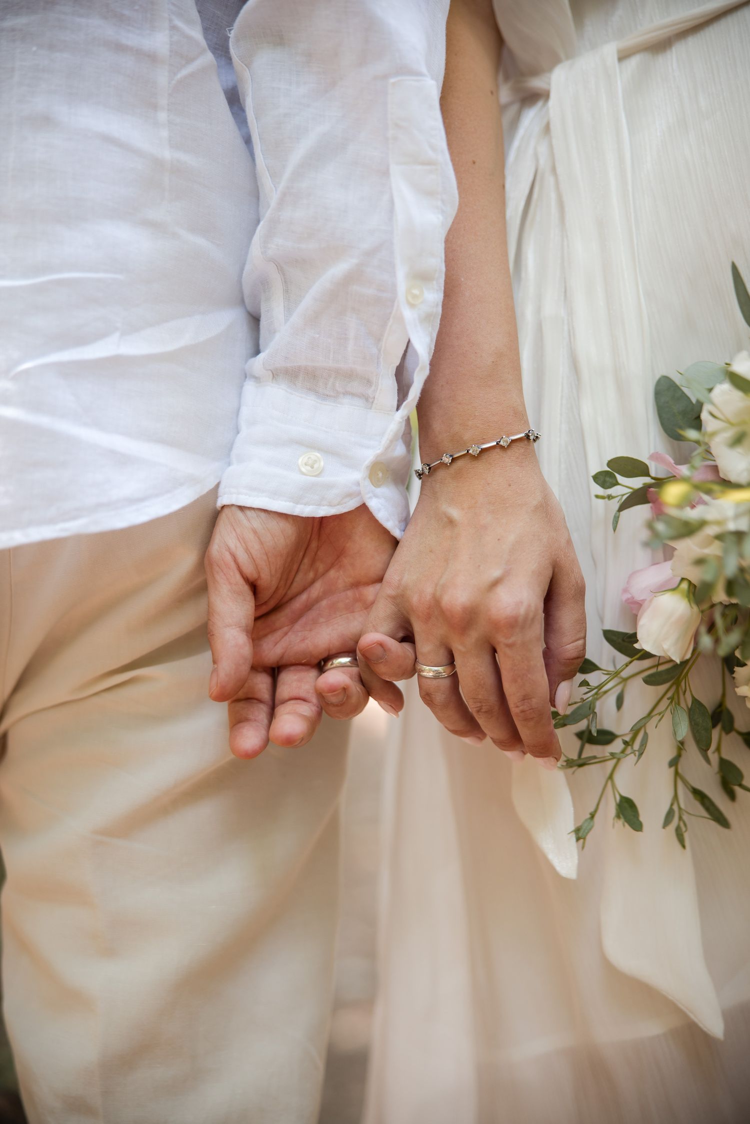 Close-up of two hands clasping together showing wedding rings against white clothing and greenery. Detail shot from bridal couple portraits