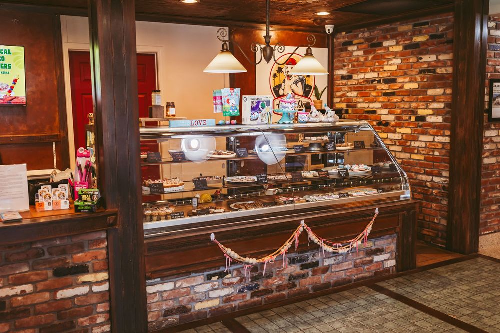 A rustic bakery display case with curved glass sits against an exposed brick wall under pendant lighting.