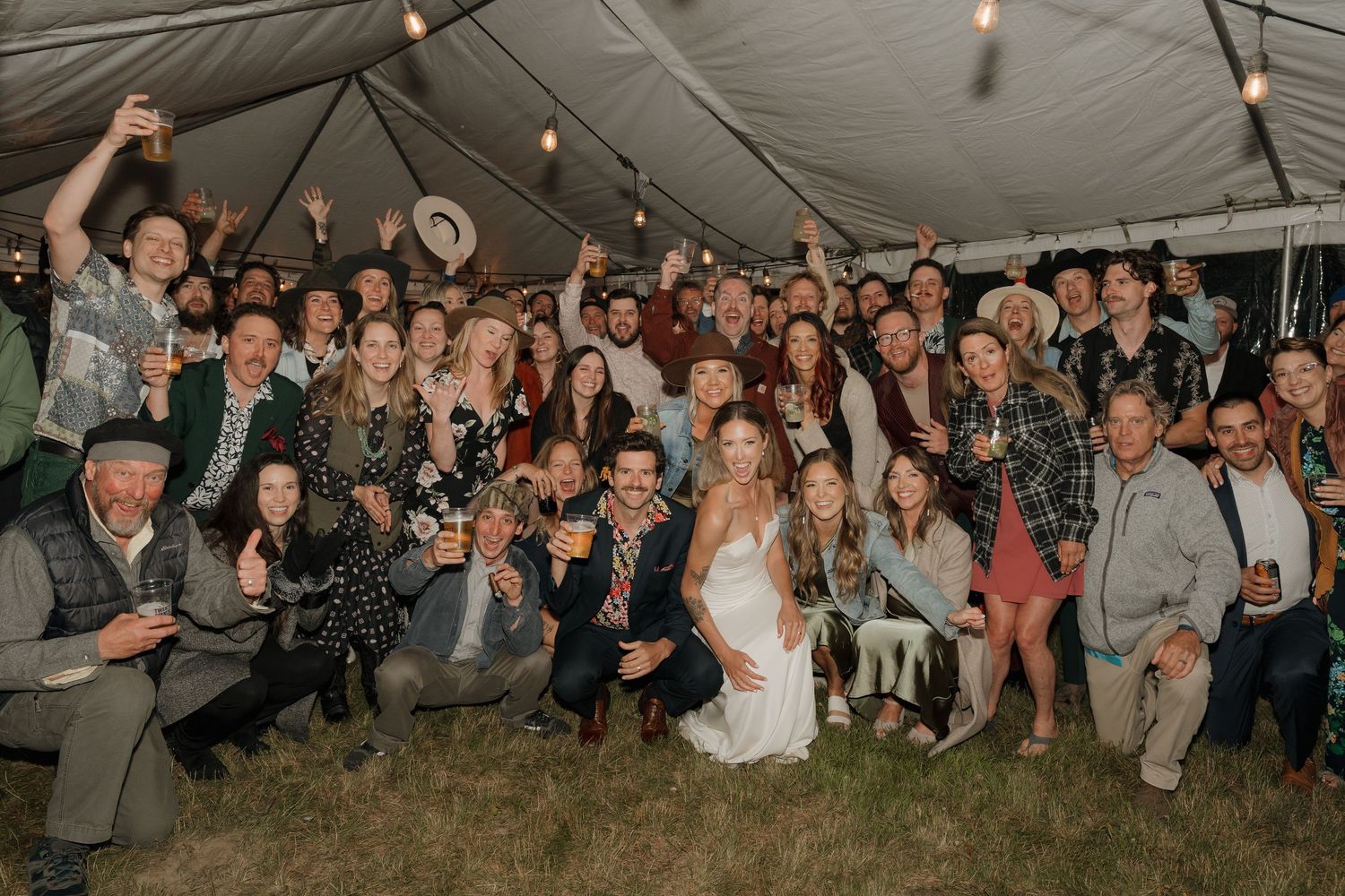 Wedding guests gather under a tent for a celebration during an outdoor evening reception.