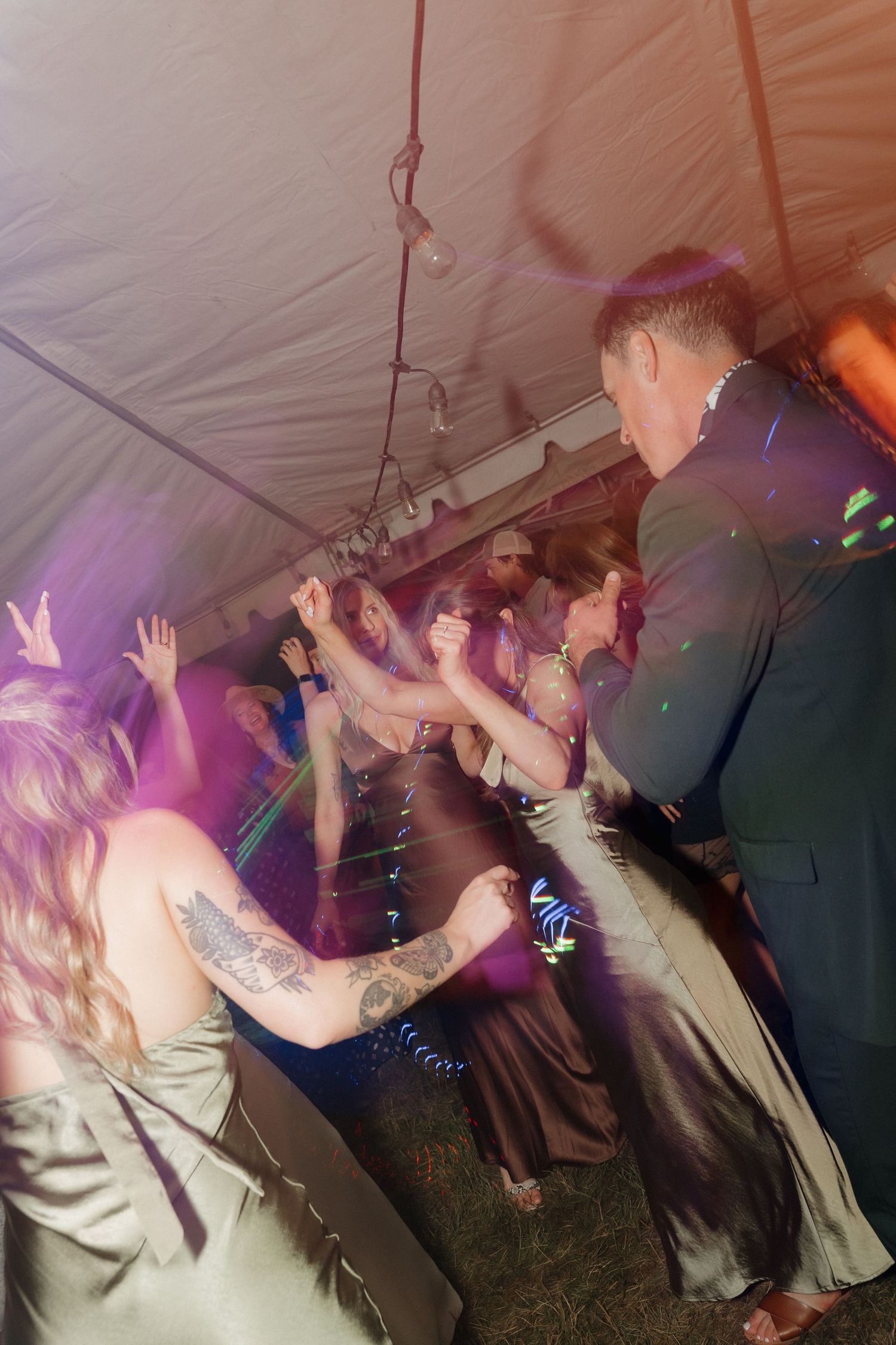 People dancing and raising their hands under a white tent at a party with purple lighting and festive atmosphere.