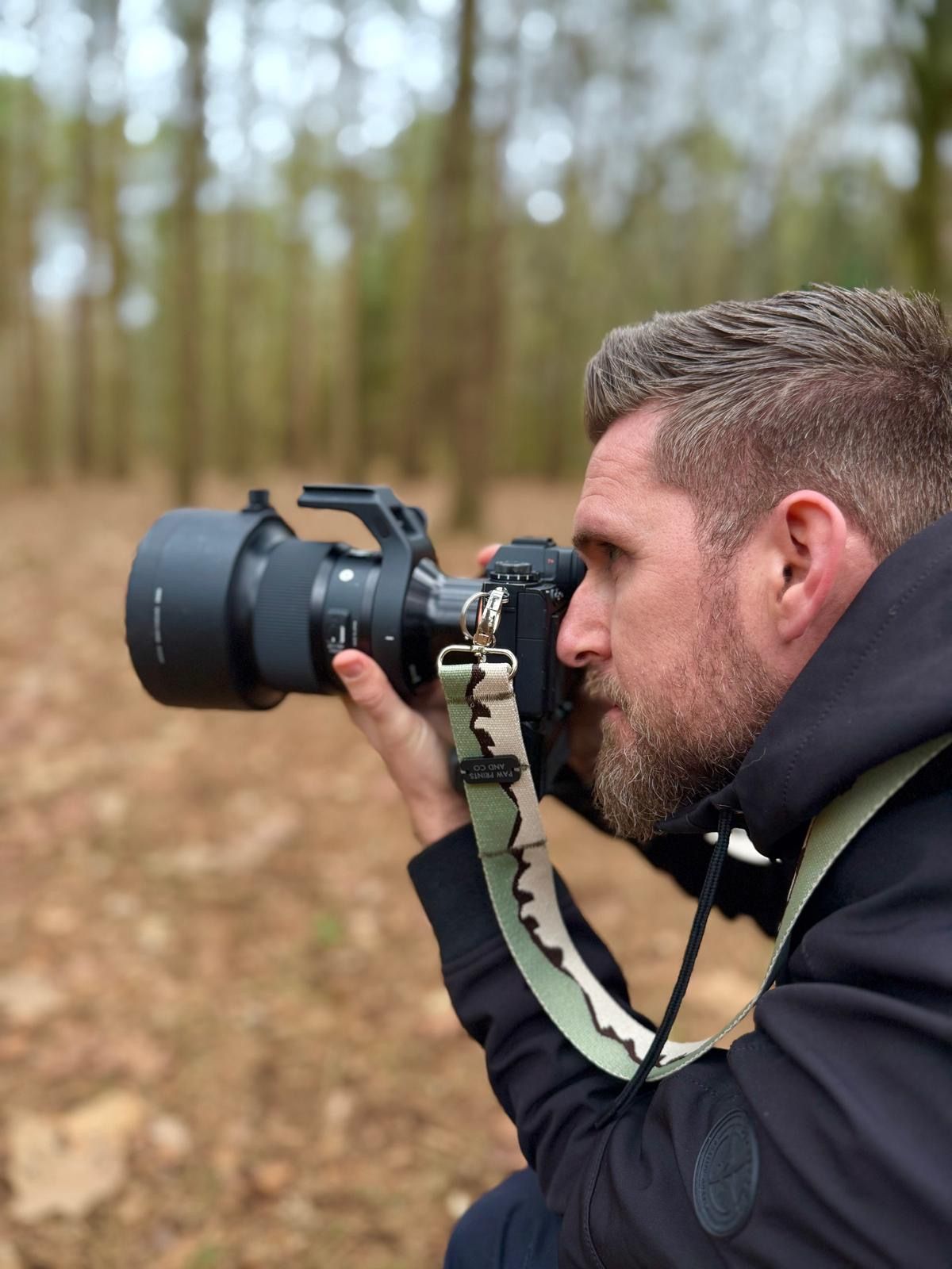 A photographer in dark clothing uses a professional camera with telephoto lens in a wooded setting.