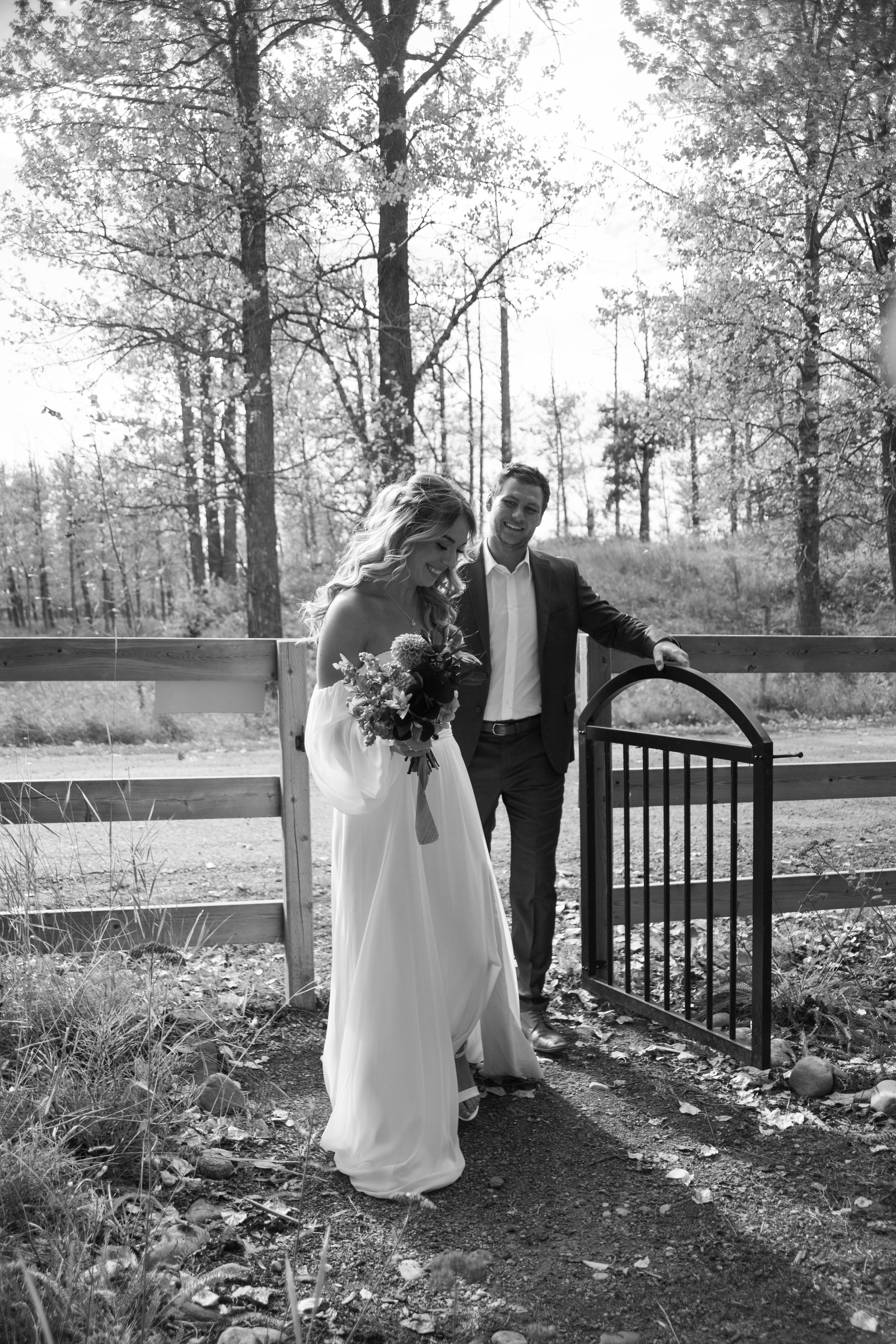Black and white wedding photo of a couple standing by a decorative gate in a wooded setting.