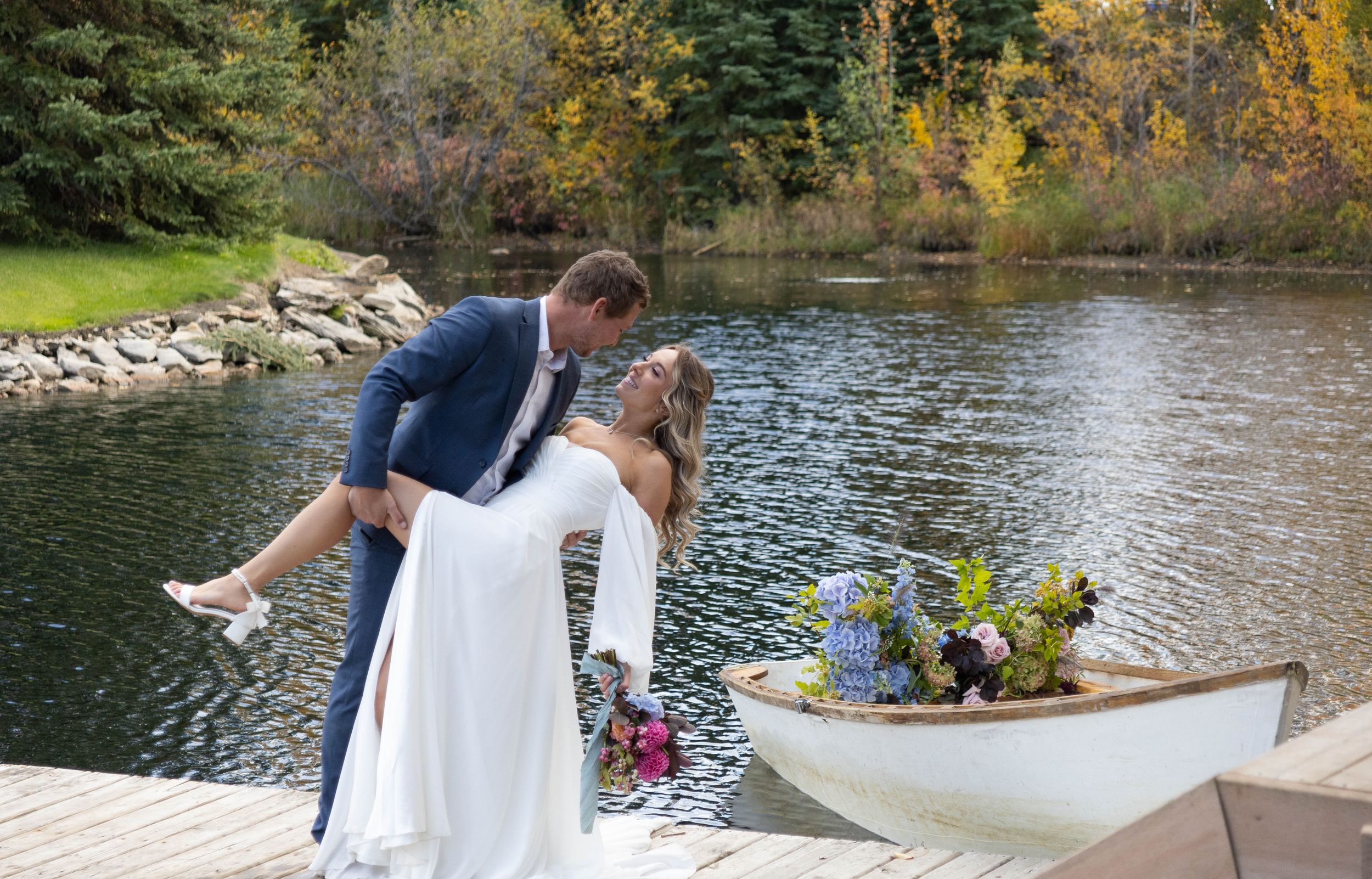 Wedding couple posing playfully by a lake with a white boat and autumn foliage in the background.