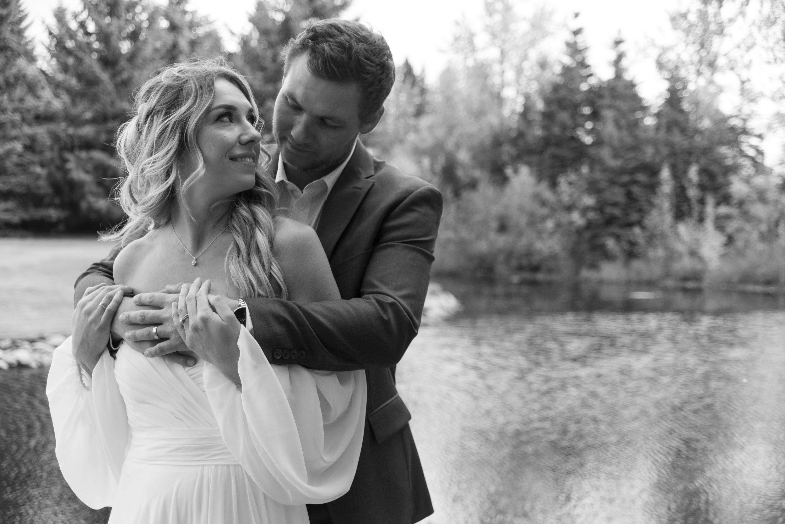 A joyful wedding couple embracing by a lake with evergreen trees in the background.
