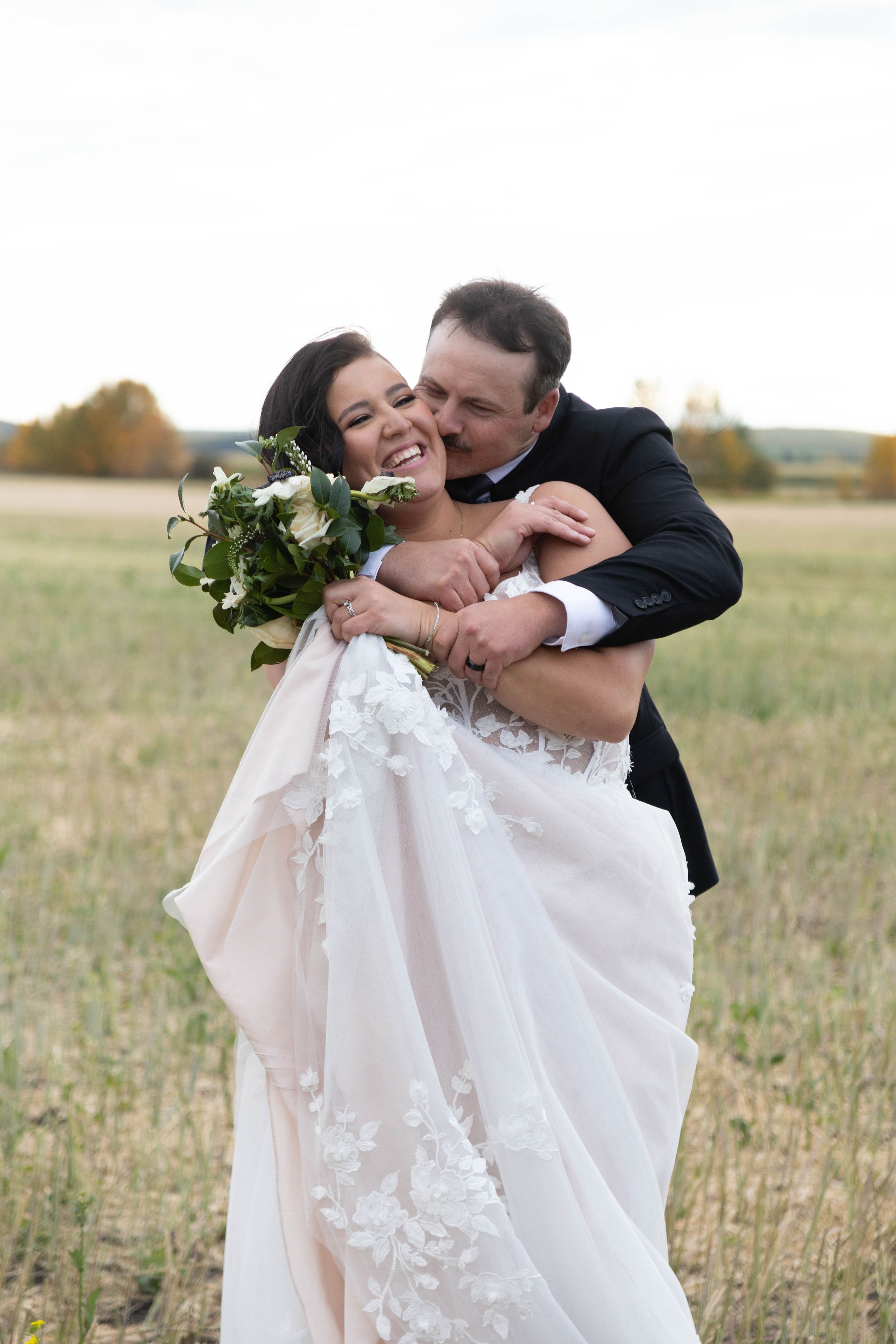 A romantic wedding portrait of a couple embracing in a field, the bride holding a white and green bouquet.