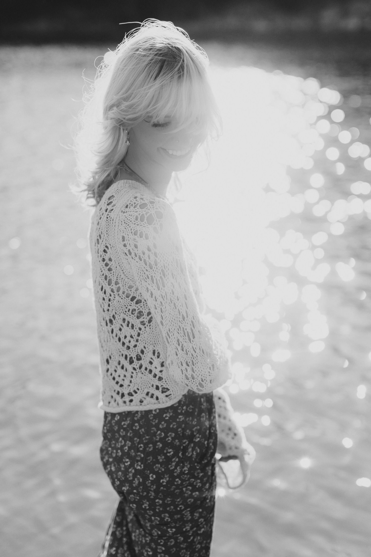 Ethereal black and white portrait of someone in a lace top and patterned skirt by the water with bokeh effects.