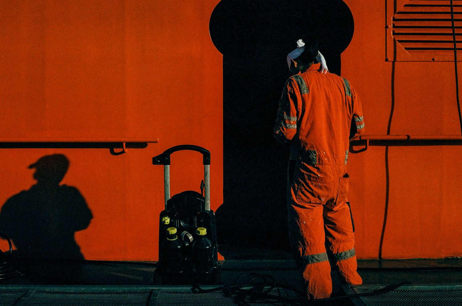 Person in orange coveralls working against bright red wall casting dramatic shadows.