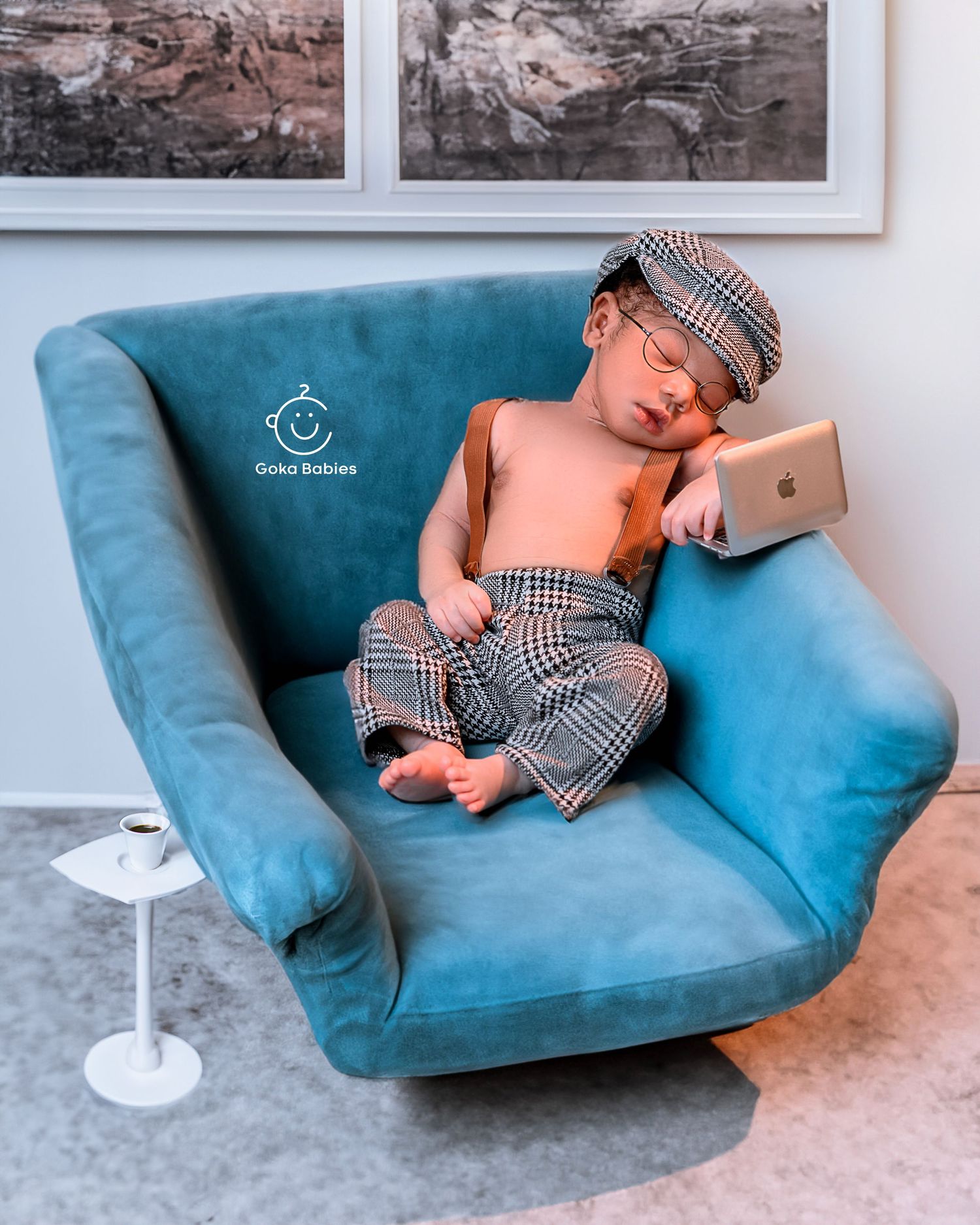 Sleeping baby in newsboy cap and plaid outfit lounging on blue velvet chair.