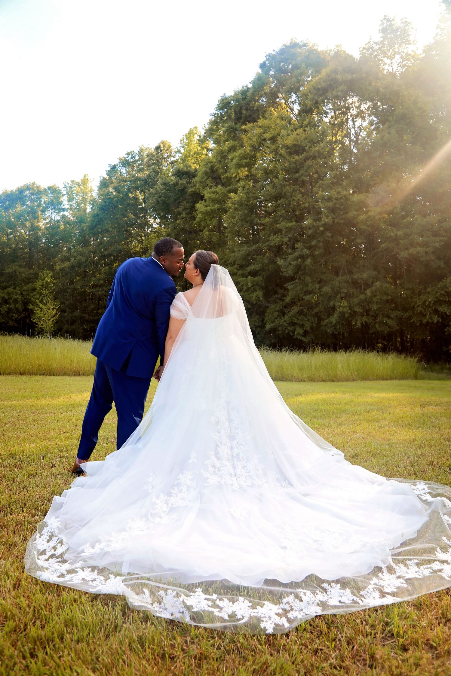 A bride and groom share an intimate moment in a sunlit field with her dramatic lace train.