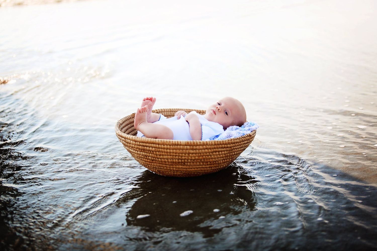 Newborn photography featuring tiny baby in woven basket floating on calm water at sunset.