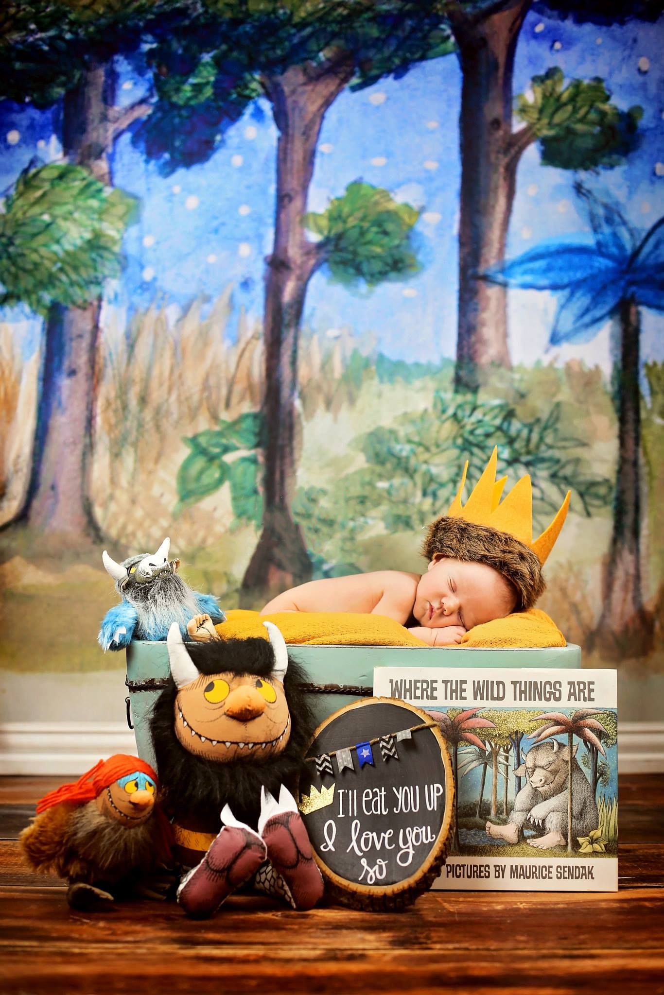 Baby sleeping beside Where the Wild Things Are book and plush toys against painted forest backdrop.