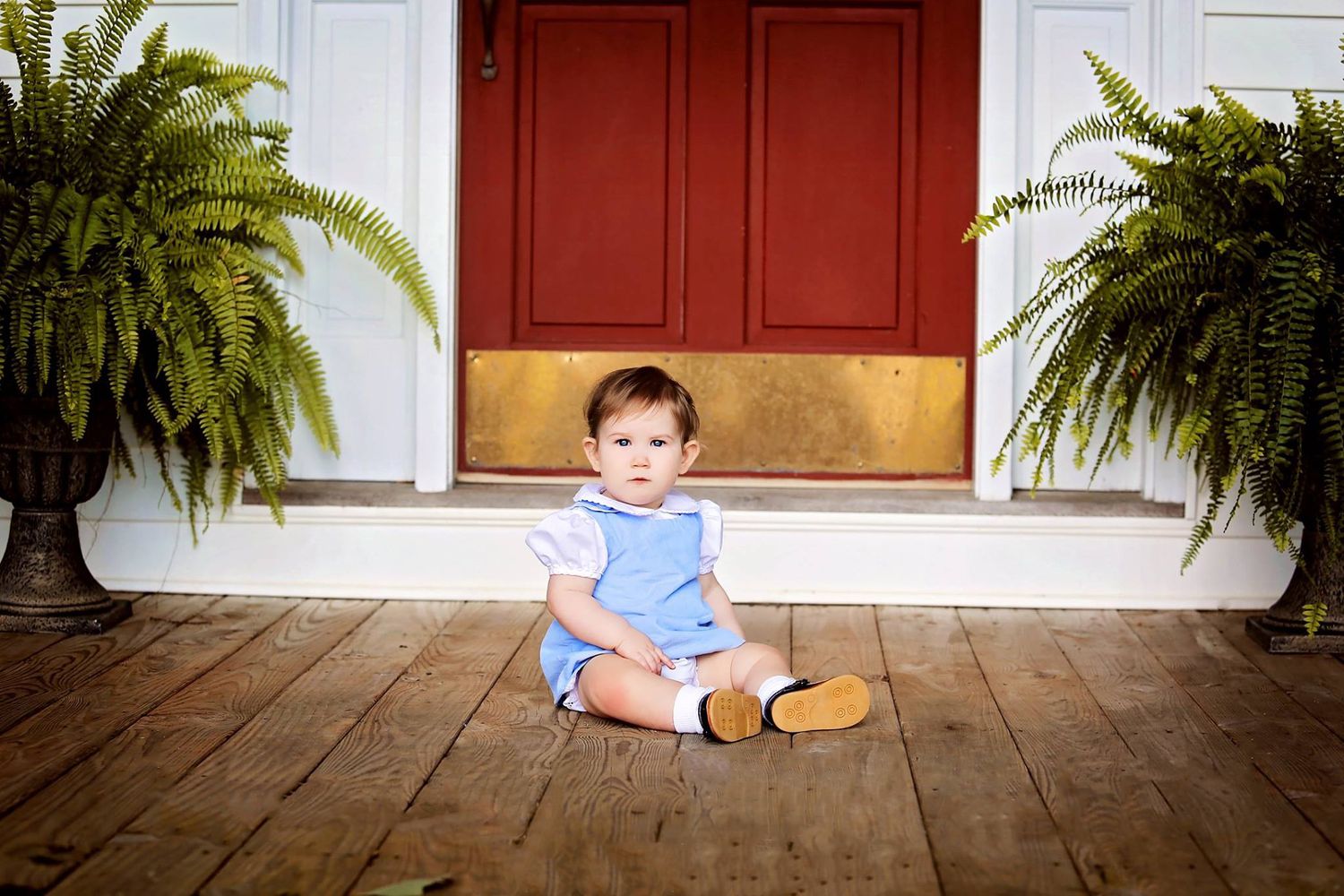 Small toddler in blue outfit sitting on porch between ferns by red door.