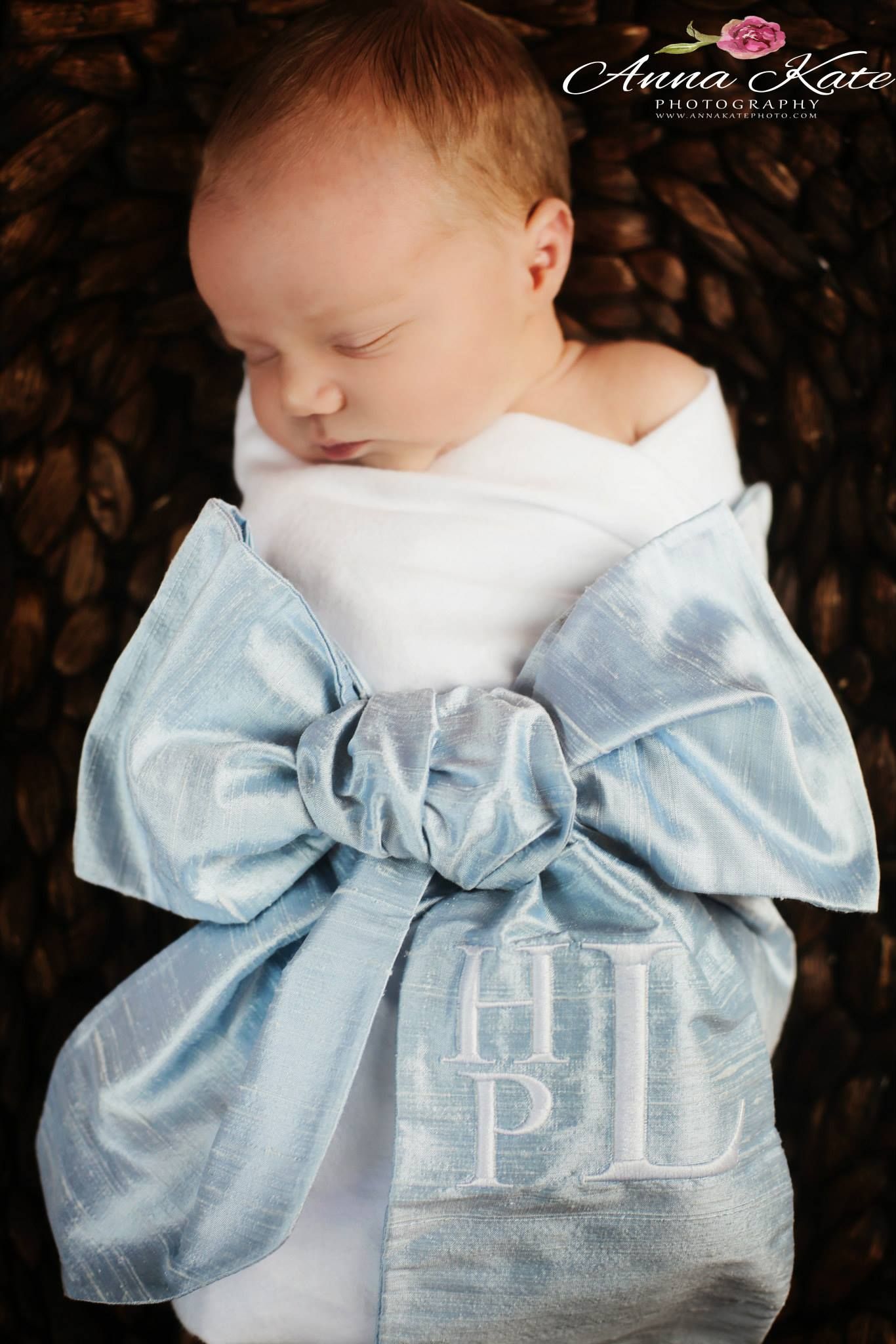 Newborn baby wrapped in a light blue bow with monogrammed initials resting on brown textured fabric.