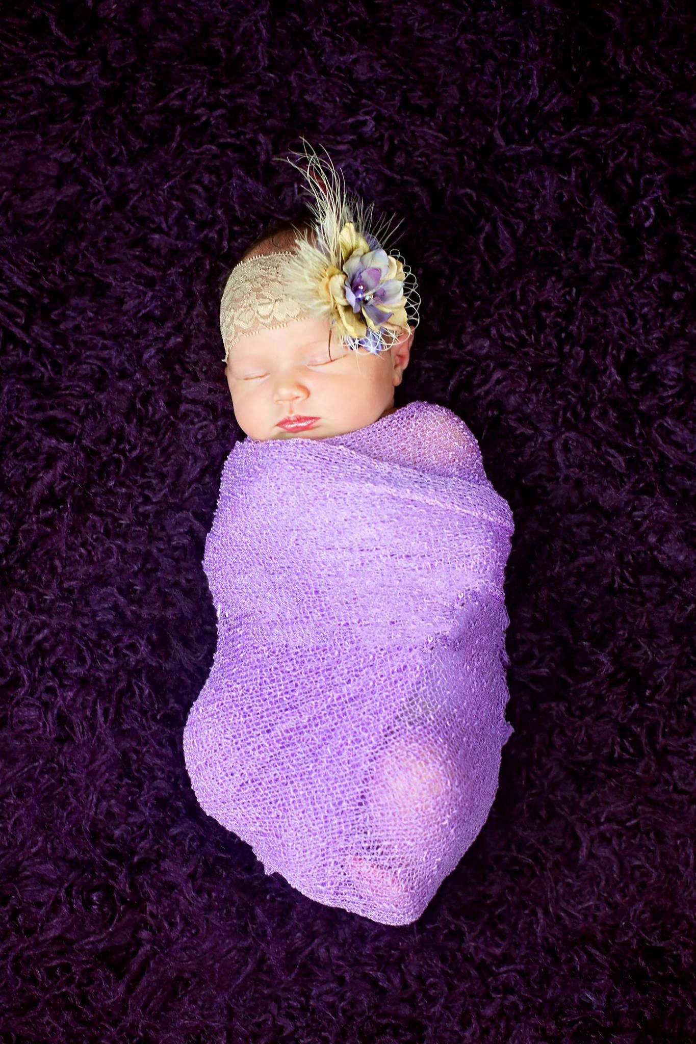 Sleeping newborn wrapped in purple swaddle with feather headband against purple background.