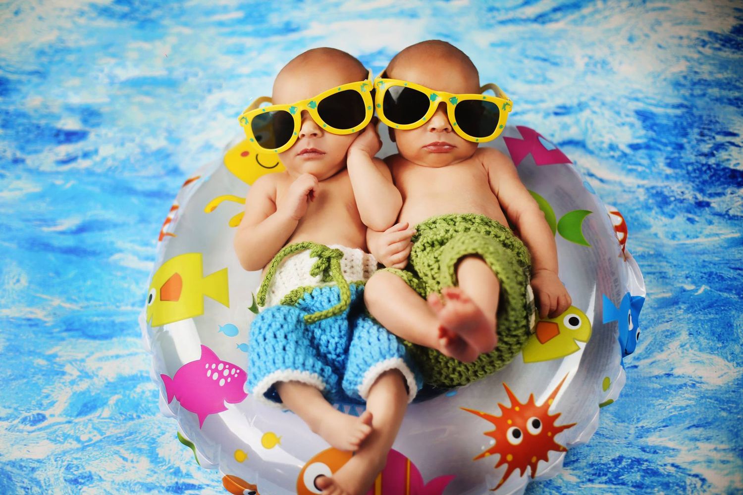 Two babies wearing sunglasses and swim shorts floating in colorful pool float.