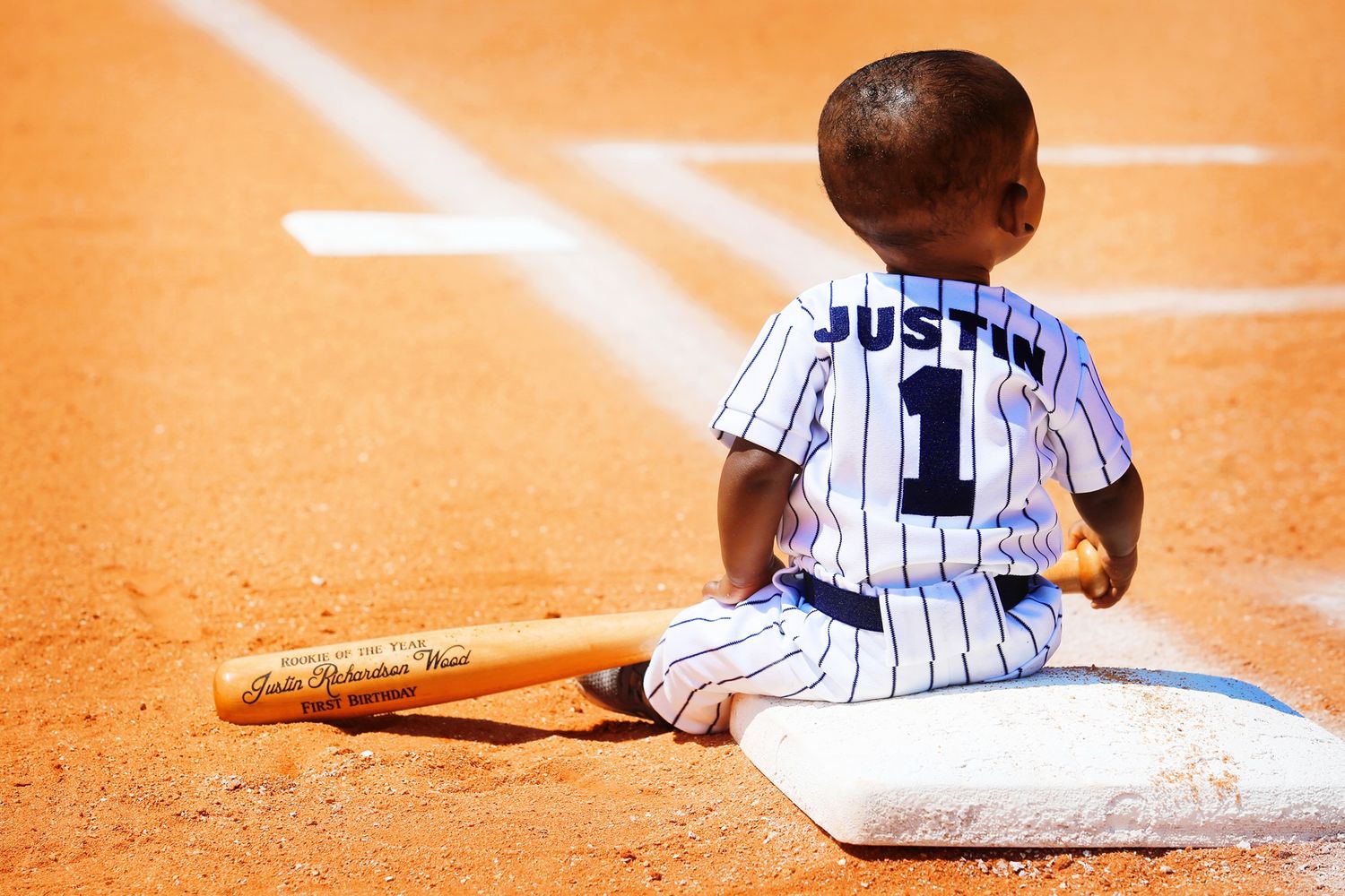 Baseball player in pinstripe uniform sitting on base with bat on dirt field.