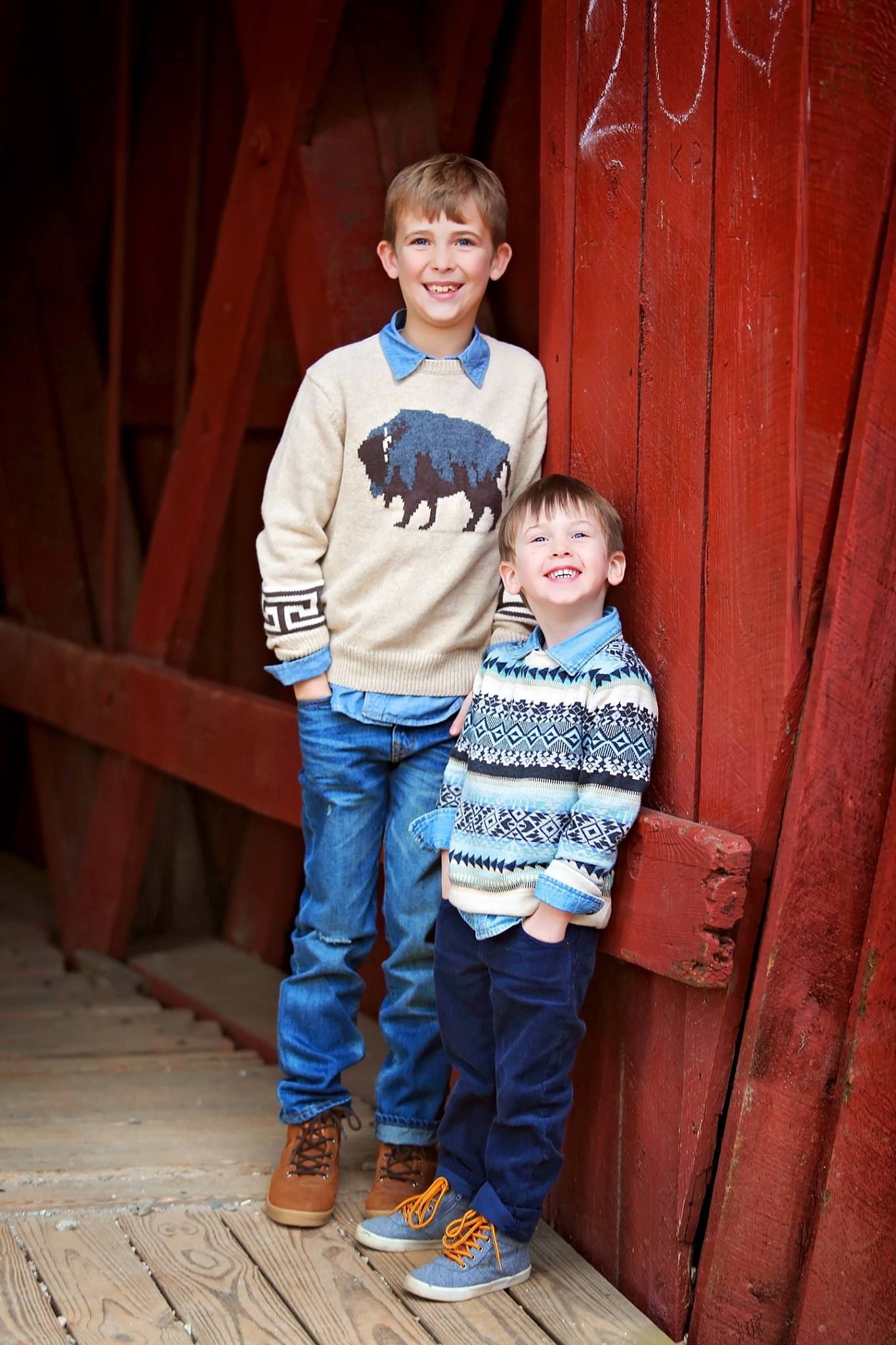 Two boys in patterned sweaters pose against red barn door.