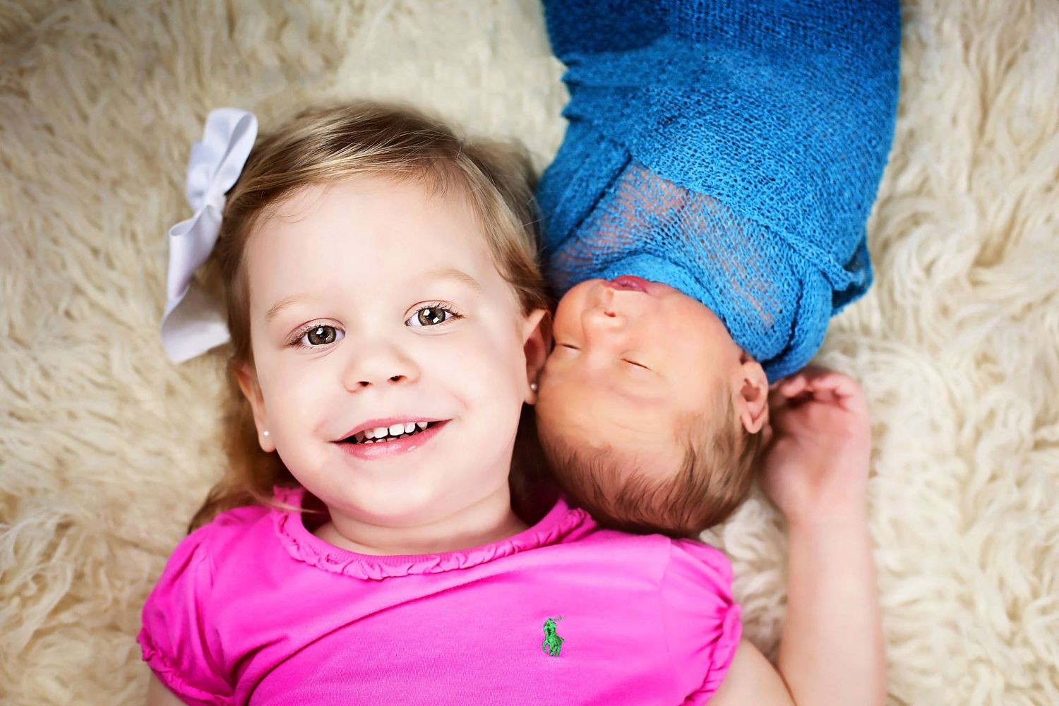 Two children laying on white fluffy rug with one in pink and one in blue.