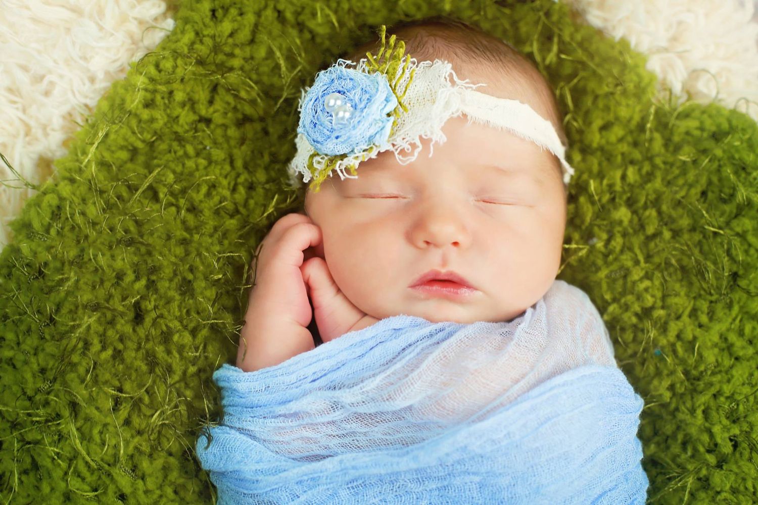 Newborn wrapped in blue fabric with floral headband sleeping on green textured backdrop.