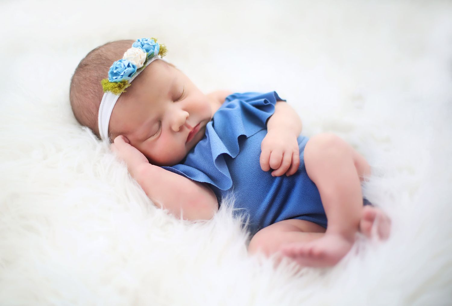Sleeping baby in blue ruffled outfit with floral headband on white fur backdrop.