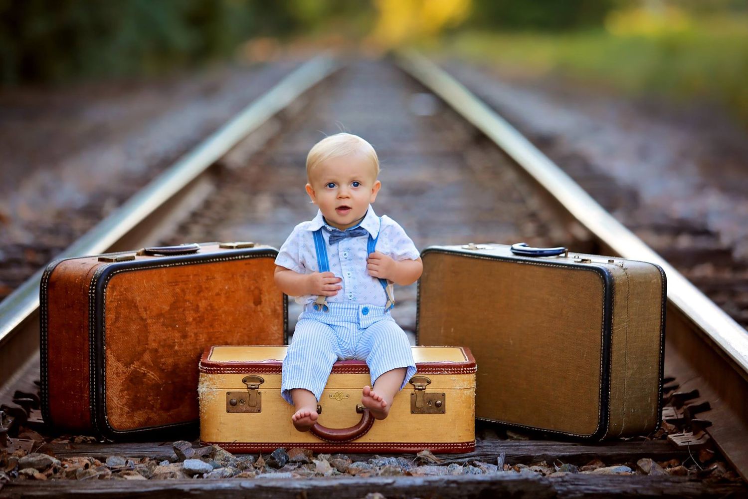 Baby in overalls sitting on vintage suitcases on train tracks.