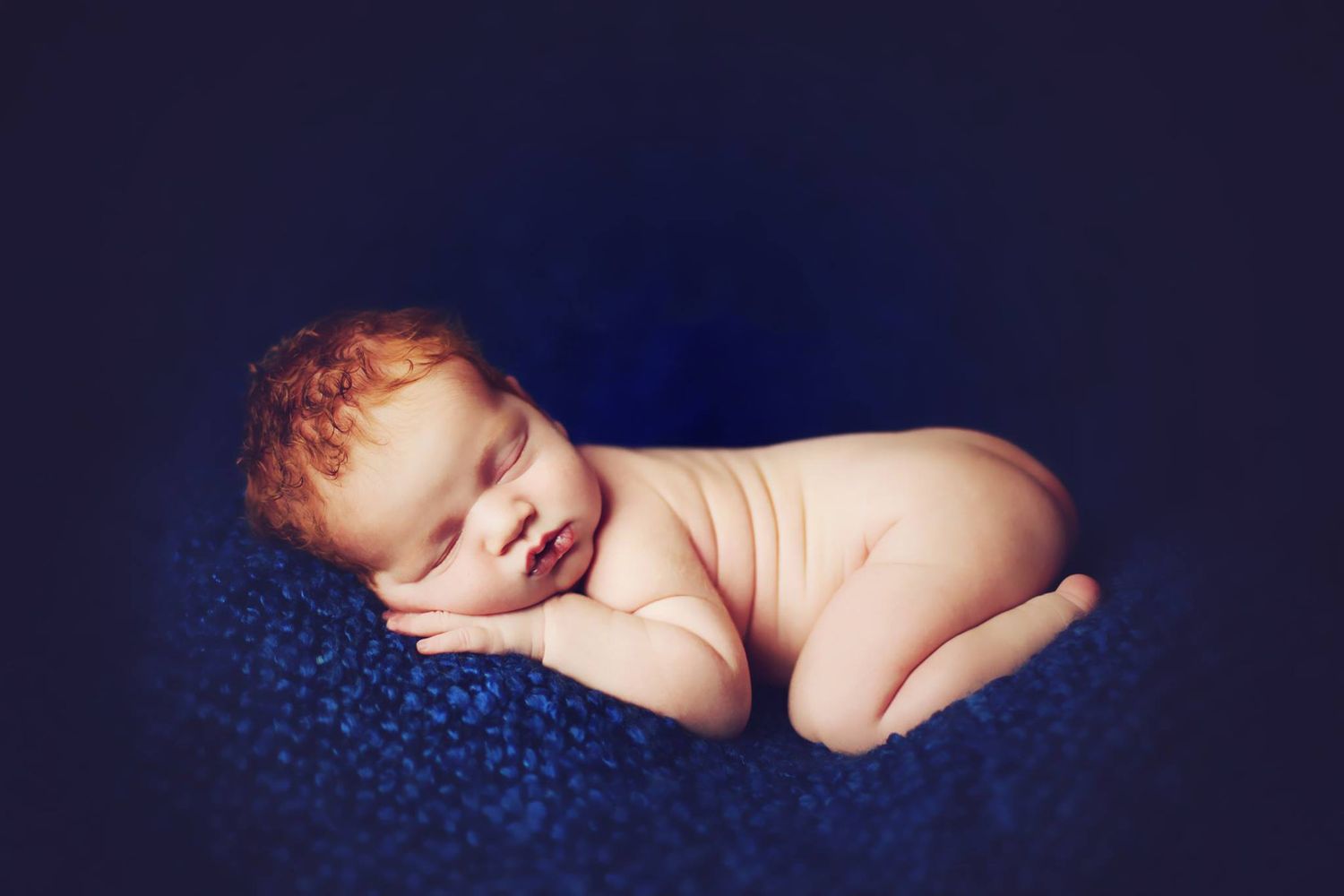 Sleeping newborn baby curled up on dark blue textured blanket.