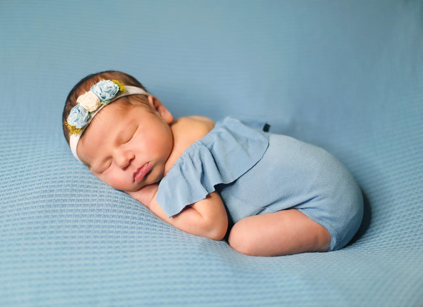 Sleeping newborn in blue outfit with floral headband on blue backdrop.
