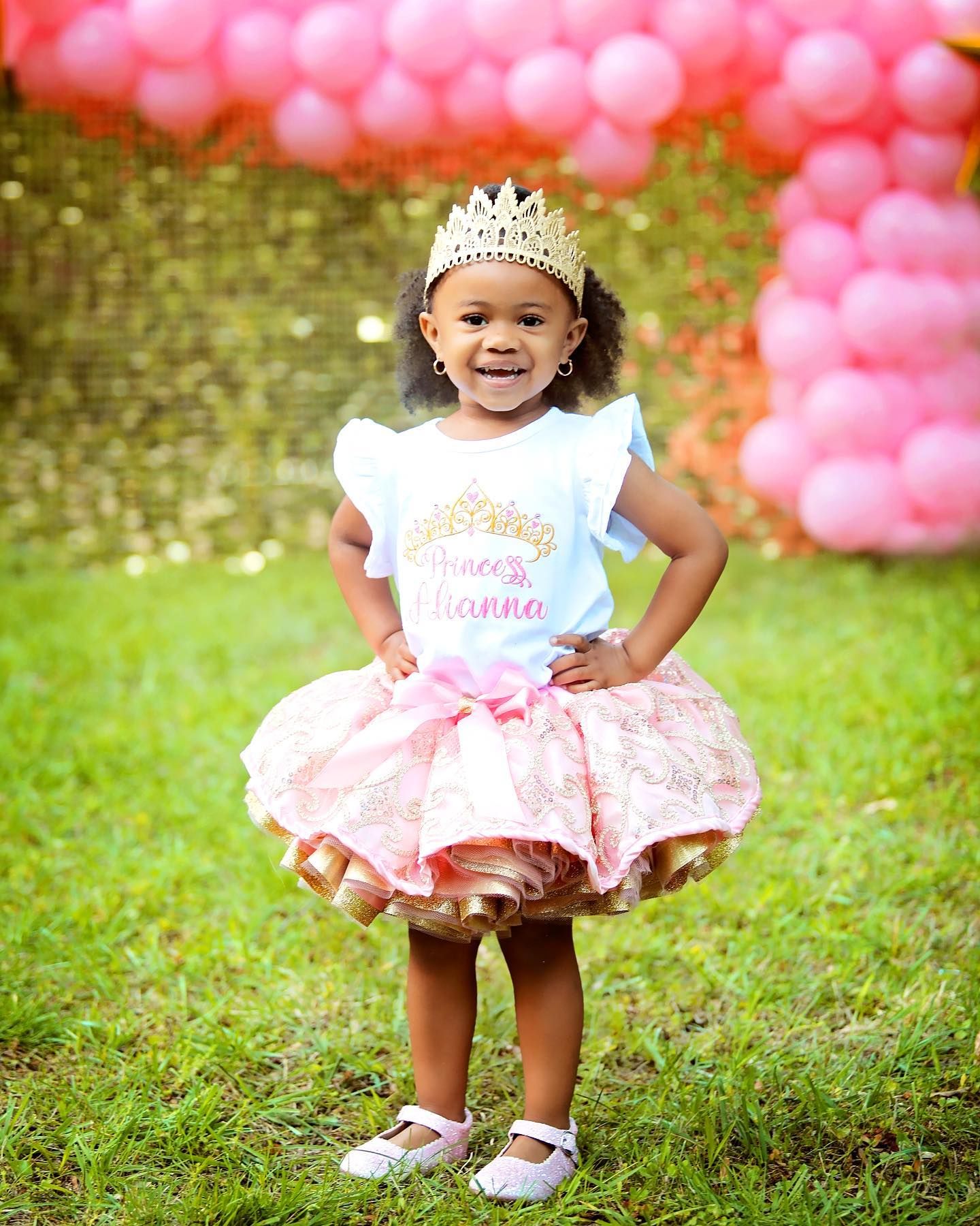 Child in princess dress and tiara poses with pink balloon backdrop.