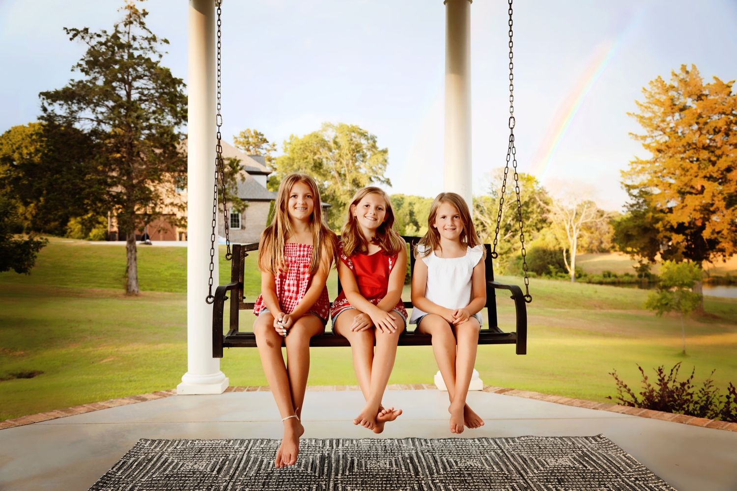 Three friends sit together on a porch swing at sunset with a rainbow visible in the background.