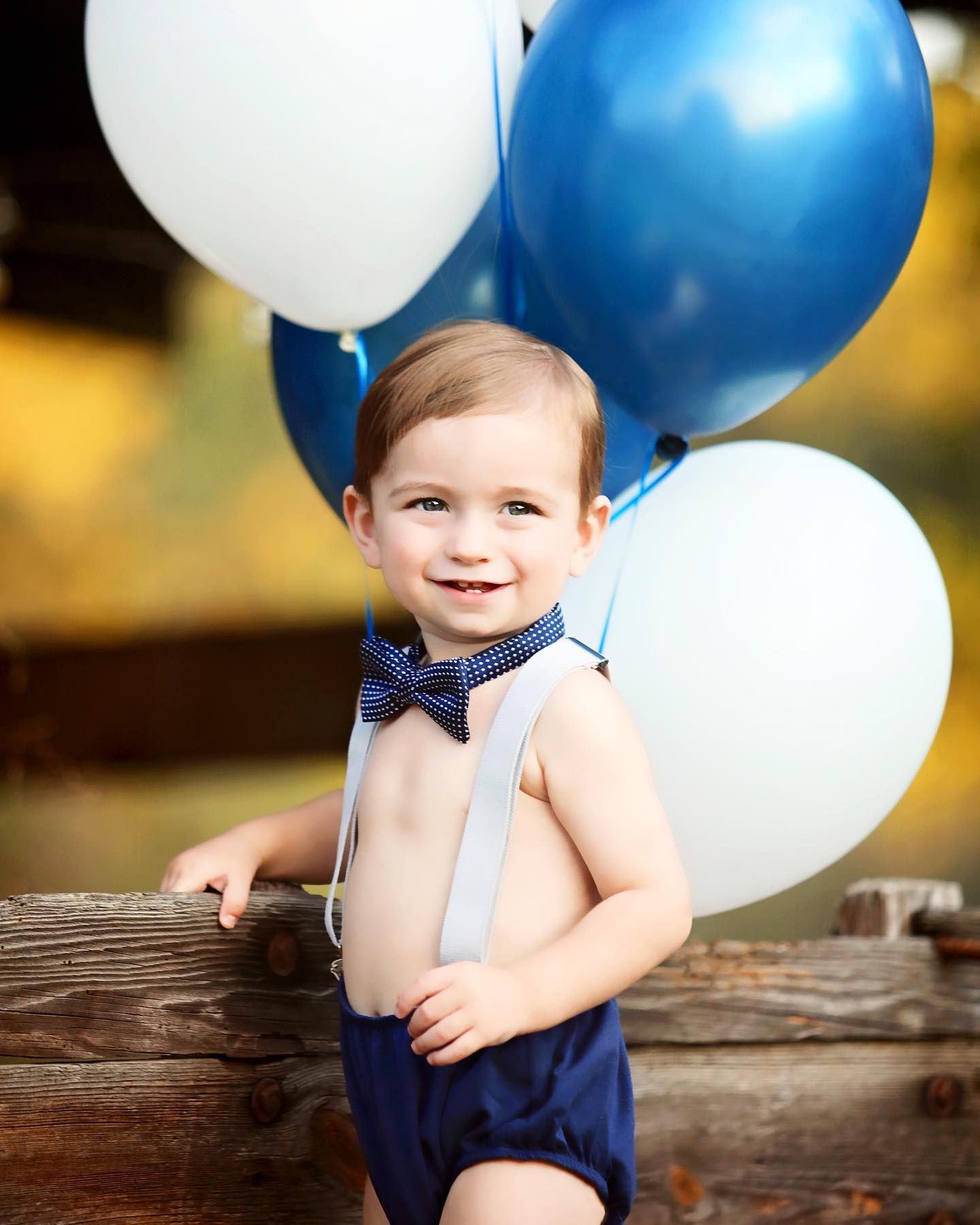 Baby in navy suspenders and bow tie poses with white and blue balloons.