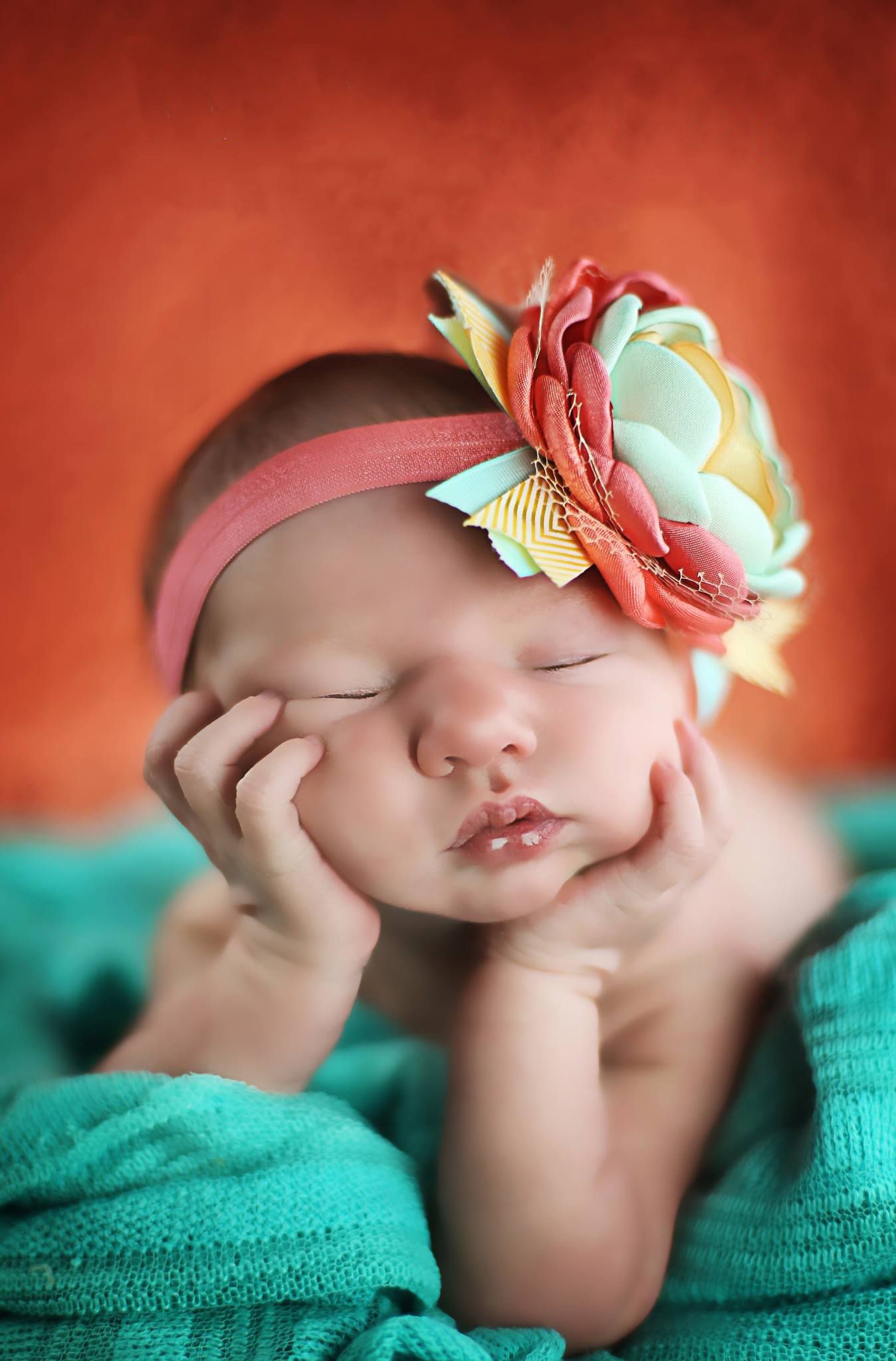 Sleeping baby rests head on hands while wearing pink floral headband and turquoise wrap in artistic portrait.