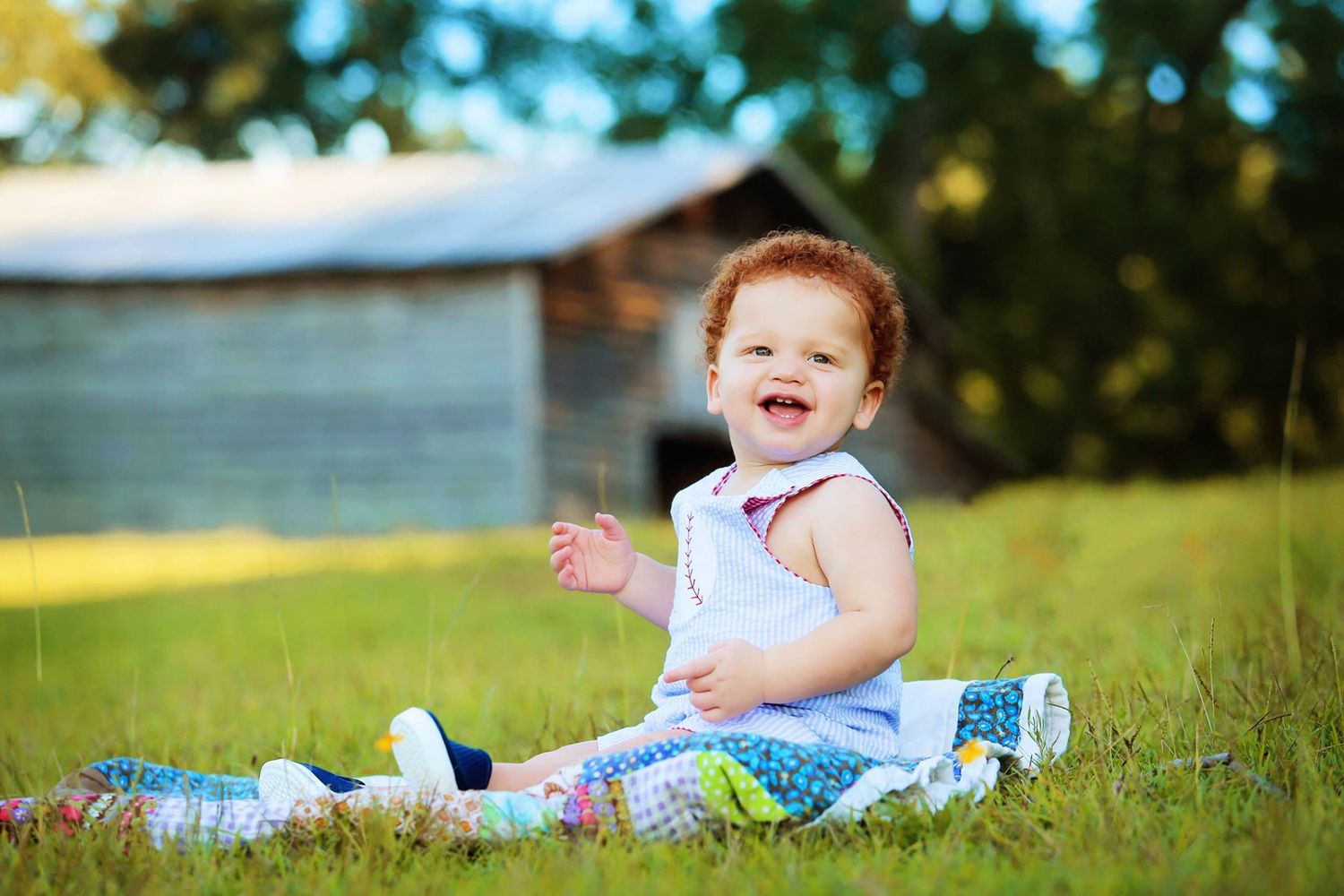 A joyful toddler in a white top sits on grass during an outdoor photo session.