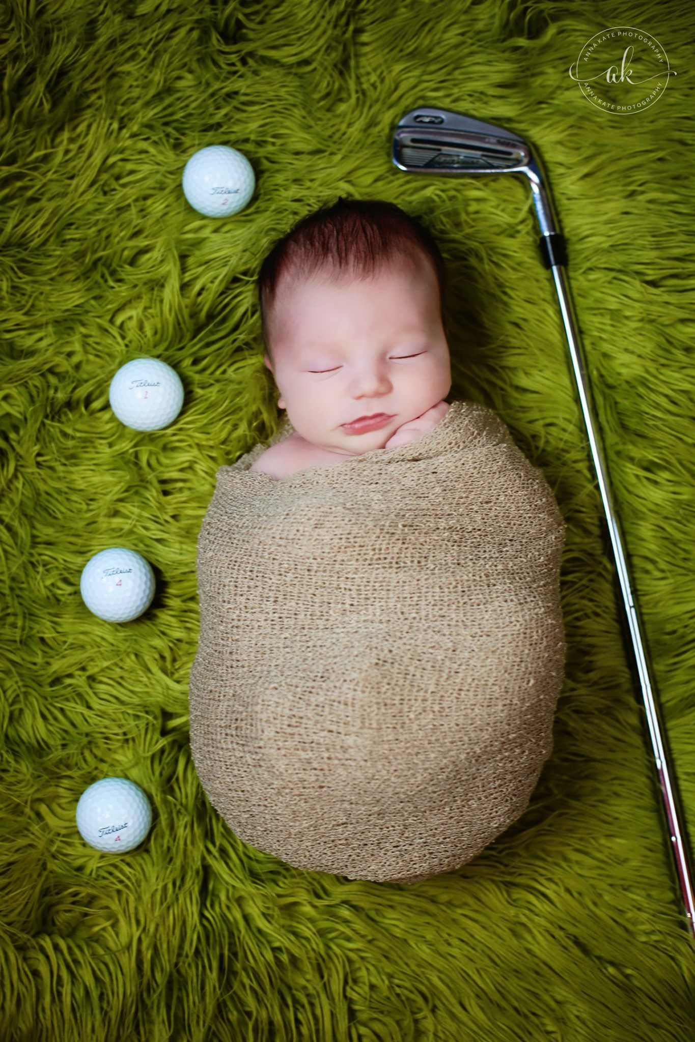Newborn wrapped in burlap lying on green carpet with golf balls and club for sports themed photo.