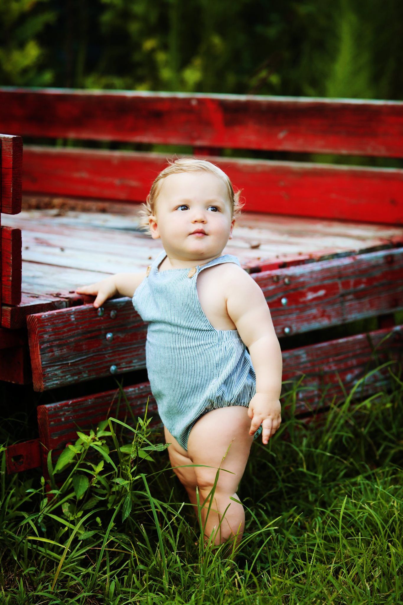 Baby in blue romper standing against red wooden bench outdoors.