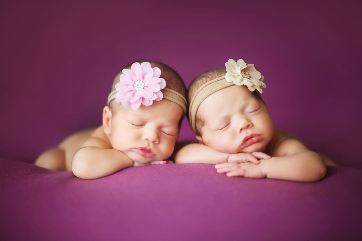 Two sleeping babies with floral headbands on purple backdrop.