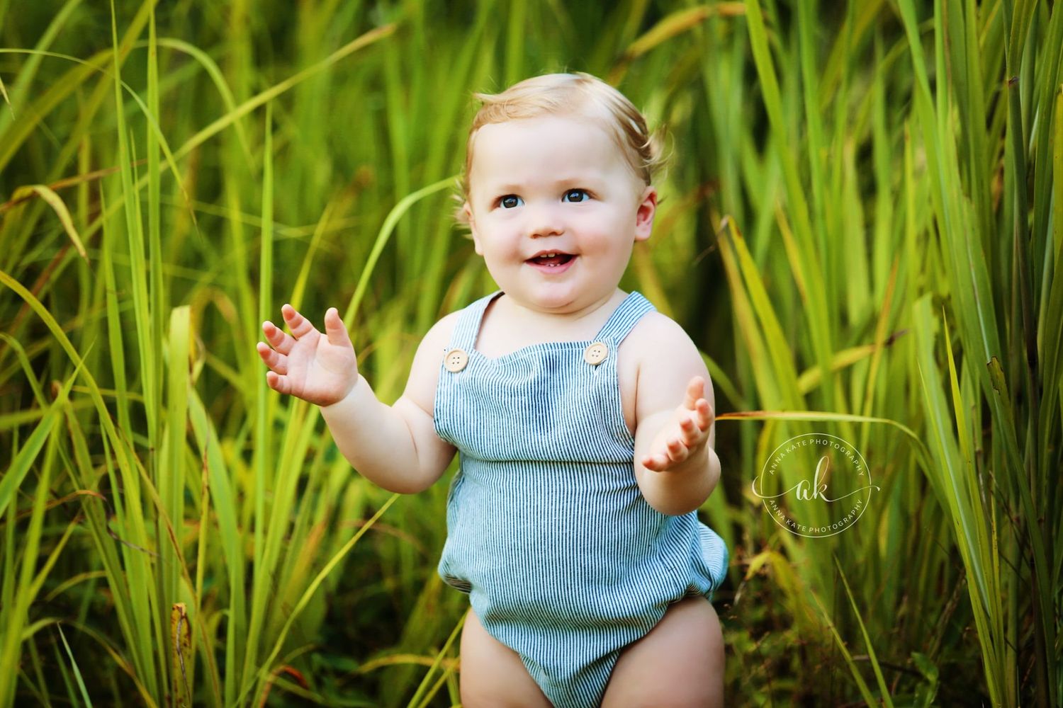 Baby in blue outfit smiling and waving while standing in tall grass.