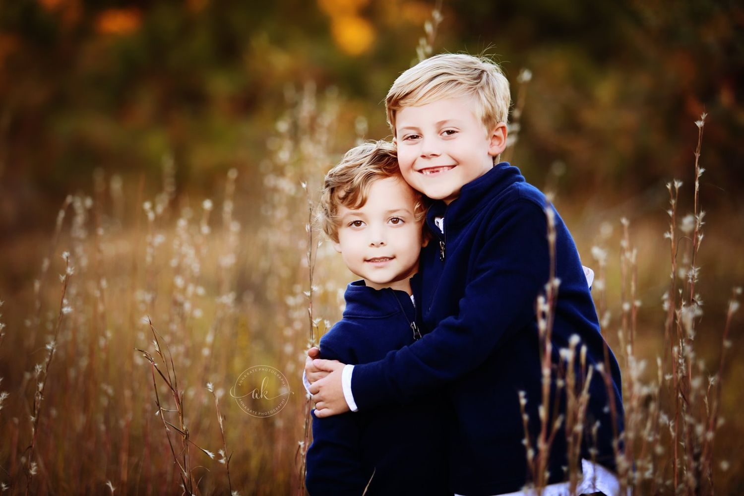 Two children in navy sweaters hugging in autumn field with tall grass.