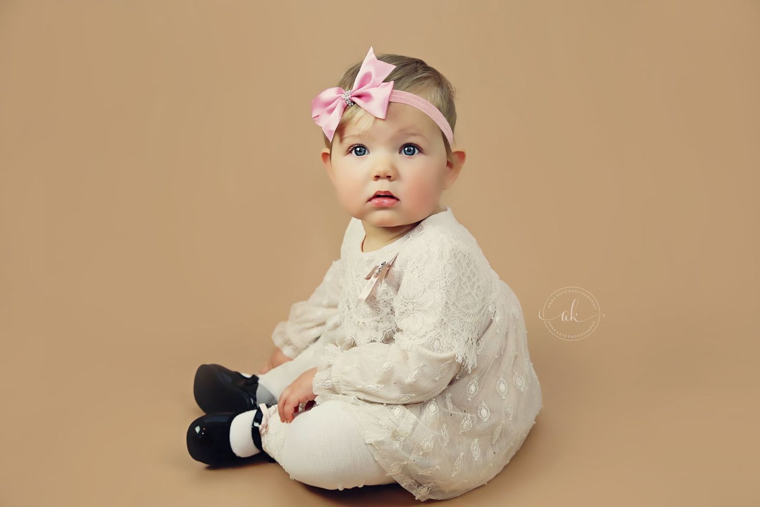 Baby in white lace dress and pink bow headband sitting against neutral backdrop.